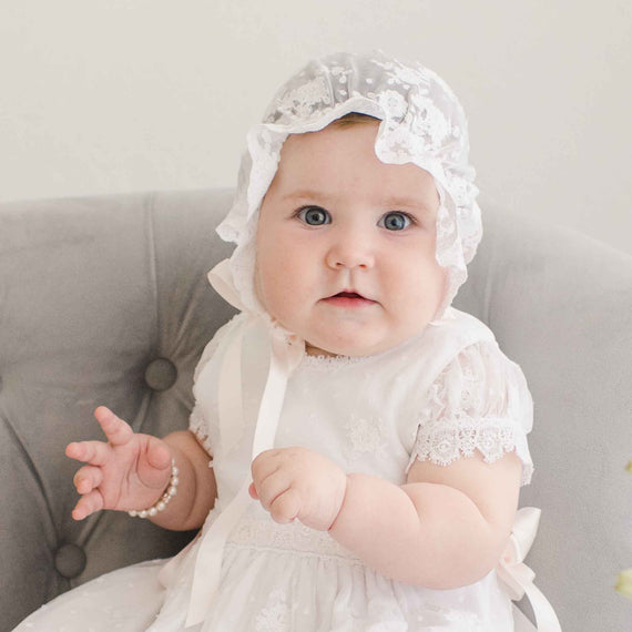 A baby with blue eyes wearing a white lace dress and the Melissa Sheer Bonnet  sits on a grey chair, looking directly at the camera with a curious expression.