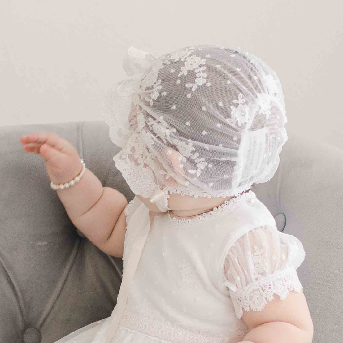 A toddler in a delicate white lace dress and matching Melissa Sheer Bonnet sits on a gray couch, playfully waving her arm. The focus is on her charming outfit, the texture of the lace.