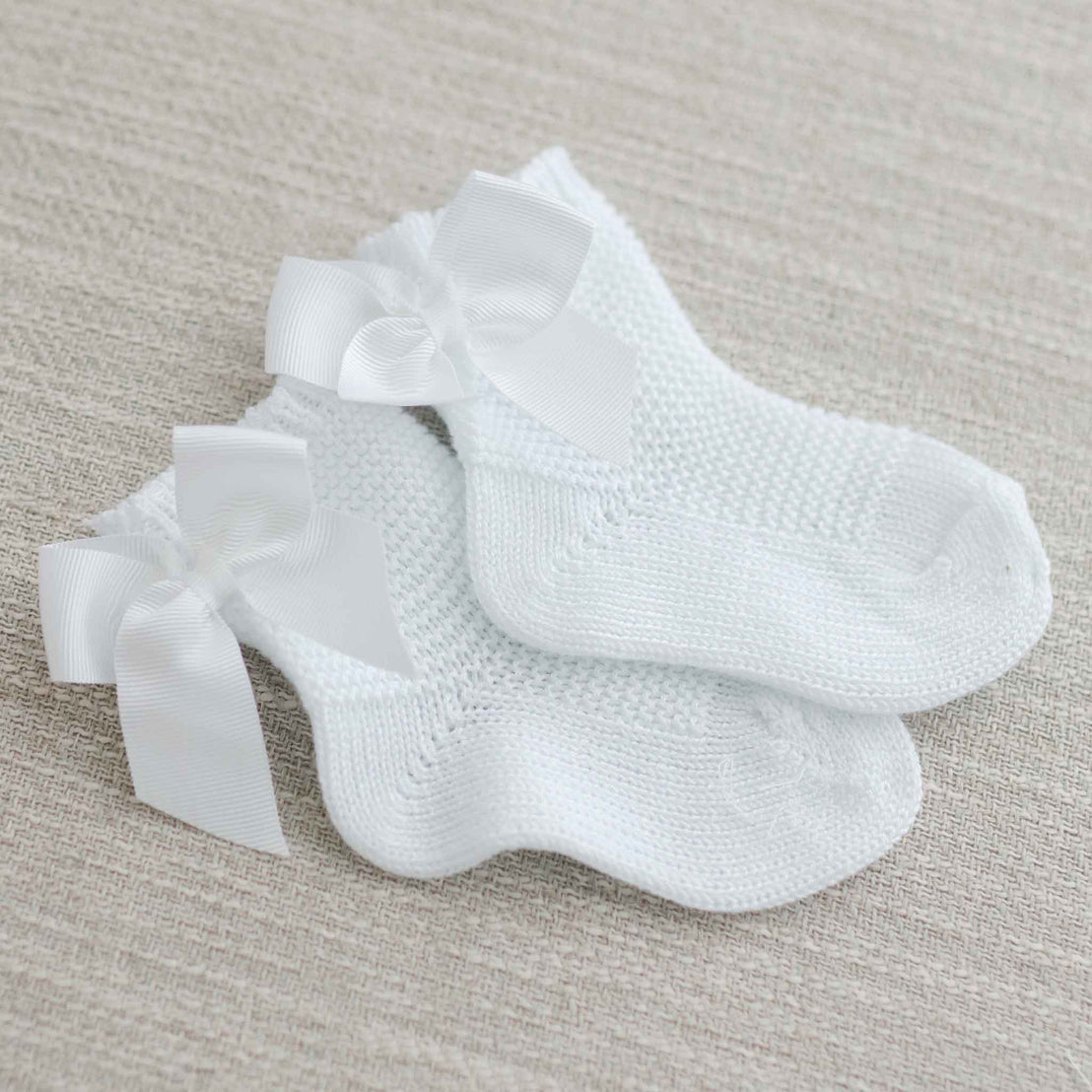 A pair of Garter Stitch Socks with Bow in white, featuring delicate lace trim and grosgrain ribbons, neatly placed on a textured beige fabric background.