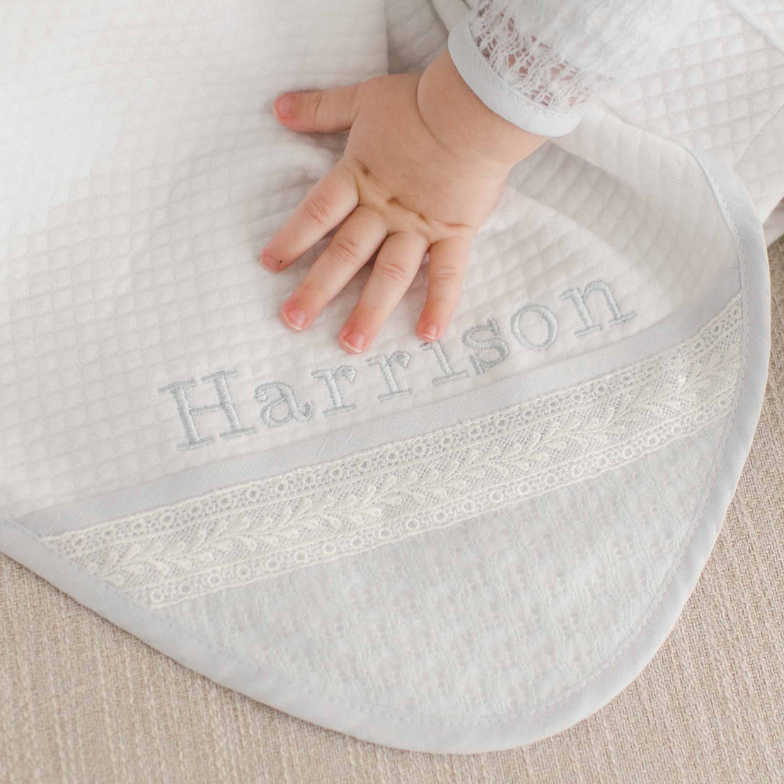 A baby's hand touches the Harrison Personalized Blanket made from plush quilted white cotton and trimmed with light blue linen. The corner of the blanket features ivory Venice lace and is embroidered in light blue thread with the name "Harrison"