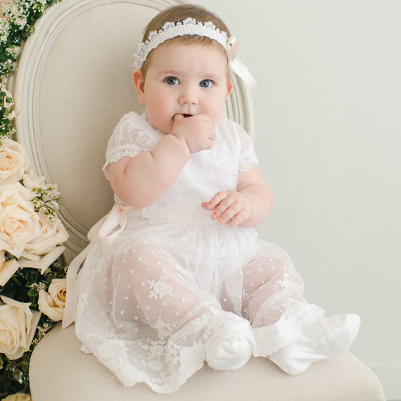 A baby dressed in a Melissa Romper Dress and headband sits on a beige chair, surrounded by cream roses, looking curiously at the camera with one hand in her mouth.