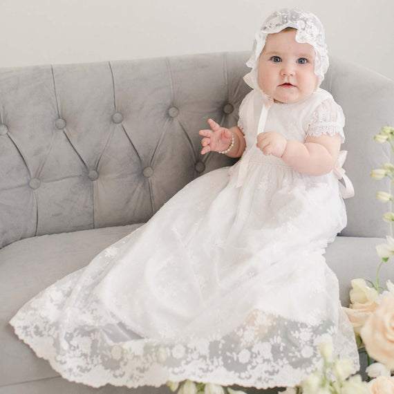 A child wearing the Melissa Christening Gown & Bonnet sits on a gray tufted sofa, surrounded by light pink roses, looking playfully at the camera.