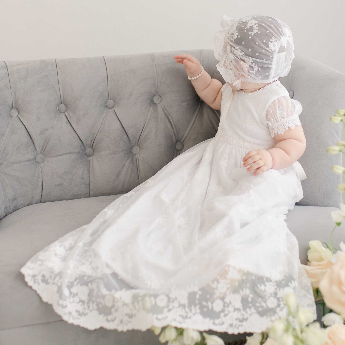 A girl dressed in a Melissa Christening Gown & Bonnet sits on a gray tufted sofa, turning her head to the side, surrounded by soft pink flowers.