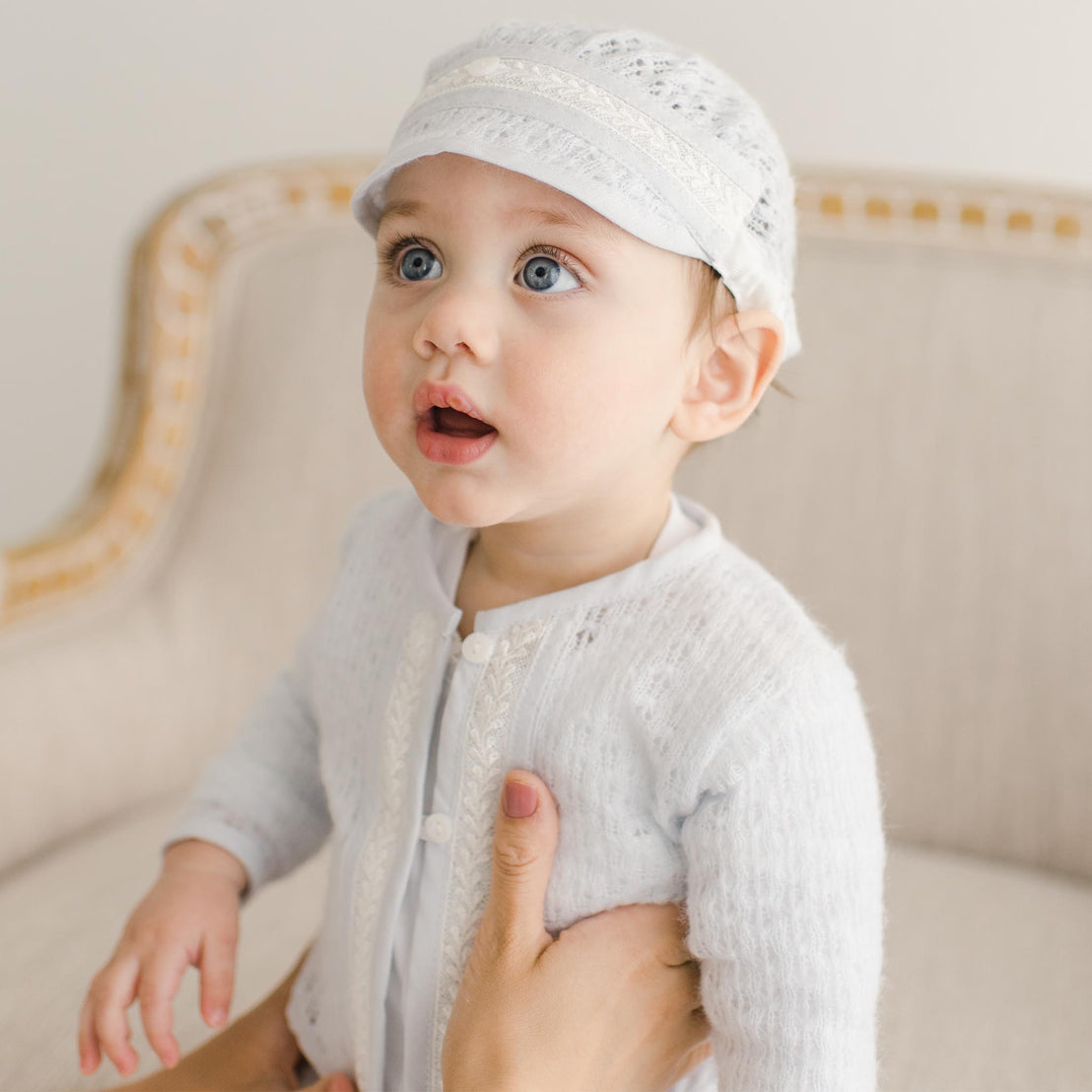 A baby boy wearing the Harrison Blue Knit Sweater and matching hat made from soft knit fabric and detailed with soft blue linen/cotton blend edging and ivory Venice lace