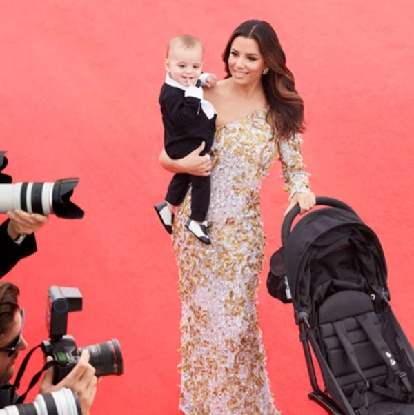 A woman in a sparkling dress smiles while holding a baby dressed in a tuxedo, standing on a red carpet. She is next to a black stroller, with photographers taking pictures around them.