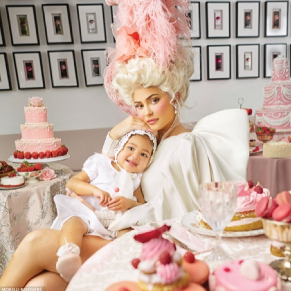 A woman dressed in an elaborate white costume with a large pink feathered headpiece holds a child on her lap. They are surrounded by pink and white desserts on a table, with framed pictures on the wall behind them.