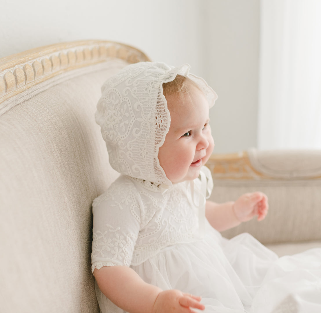 A joyful baby in a vintage white lace dress and Eliza Bonnet sits on an upscale beige sofa, smiling and looking to the side with a bright expression.