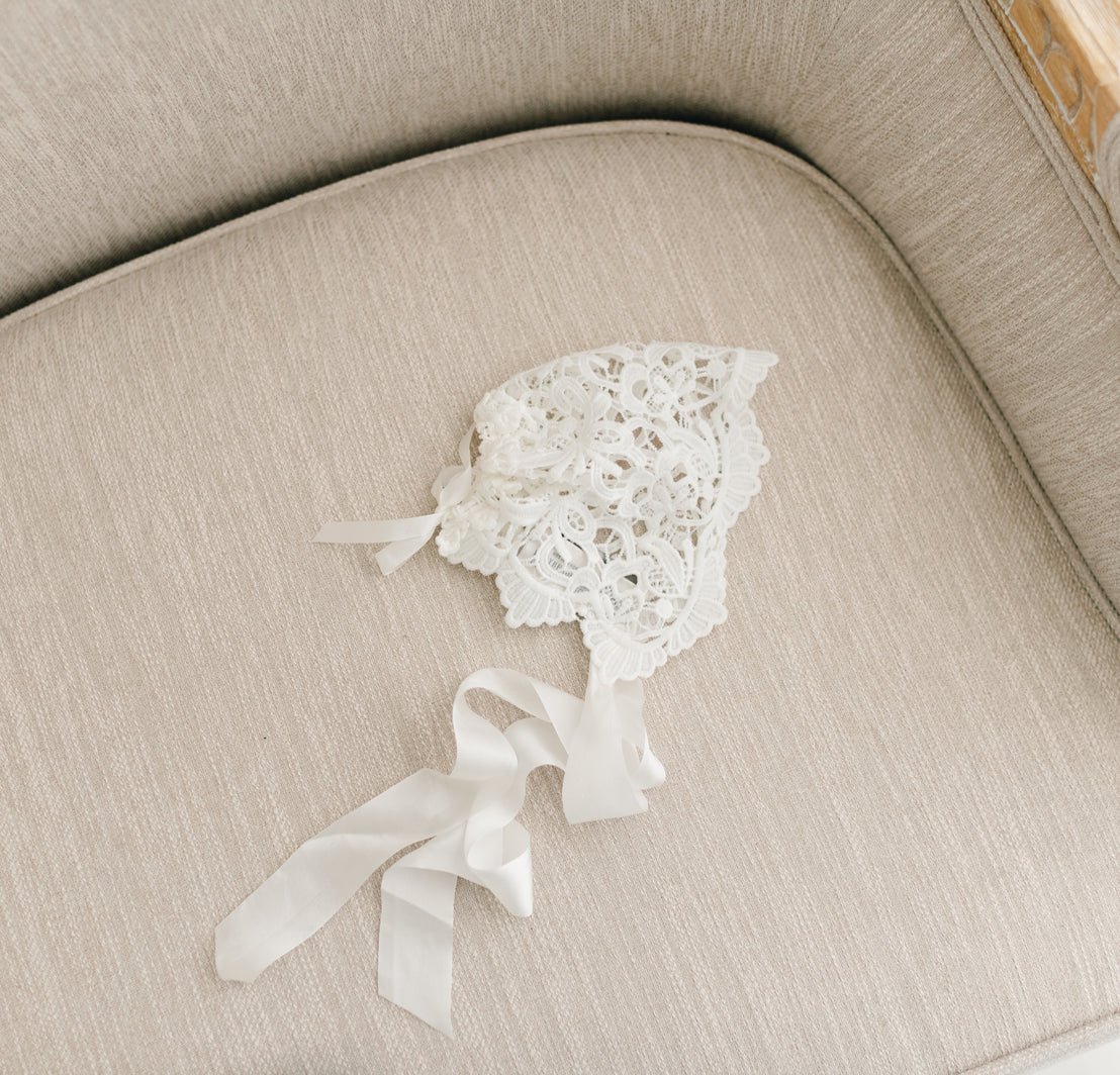 Flat lay of the Lola Lace Bonnet. The lace bonnet is designed with richly embroidered light ivory lace and cotton floral edge lace at the back.