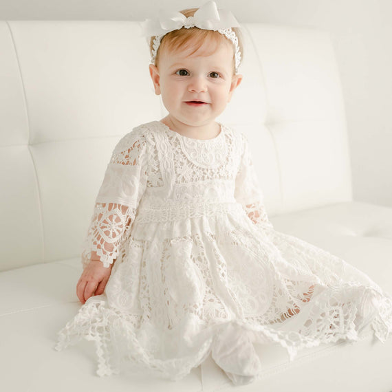 A toddler in the Adeline Lace Dress & Bloomers and matching bow headband, adorned with baby jewelry, sits on a luxury white couch, looking directly at the camera with a soft smile.