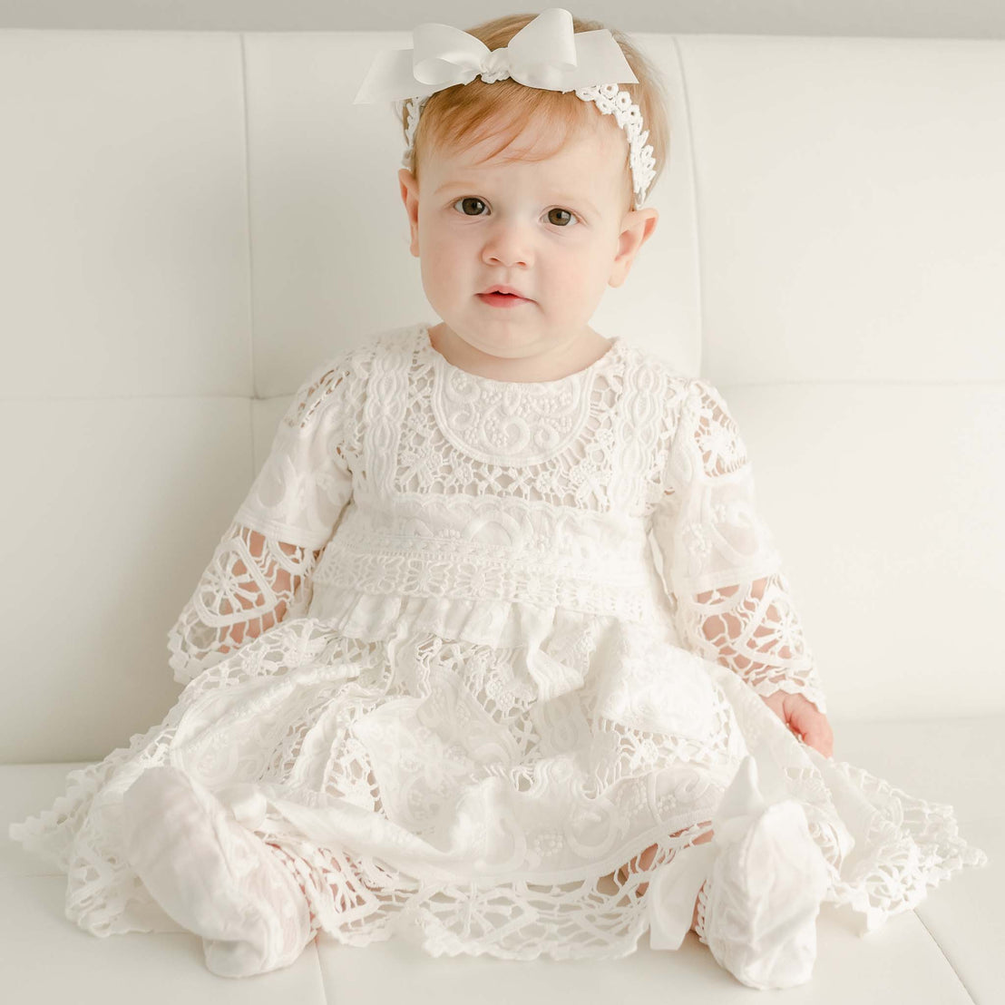 Baby girl with red hair sitting on couch wearing the Adeline lace christening dress and ribbon bow headband.