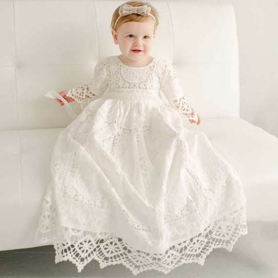 Baby girl in the Adeline Lace Cotton Christening Gown. Pictured on the couch smiling.