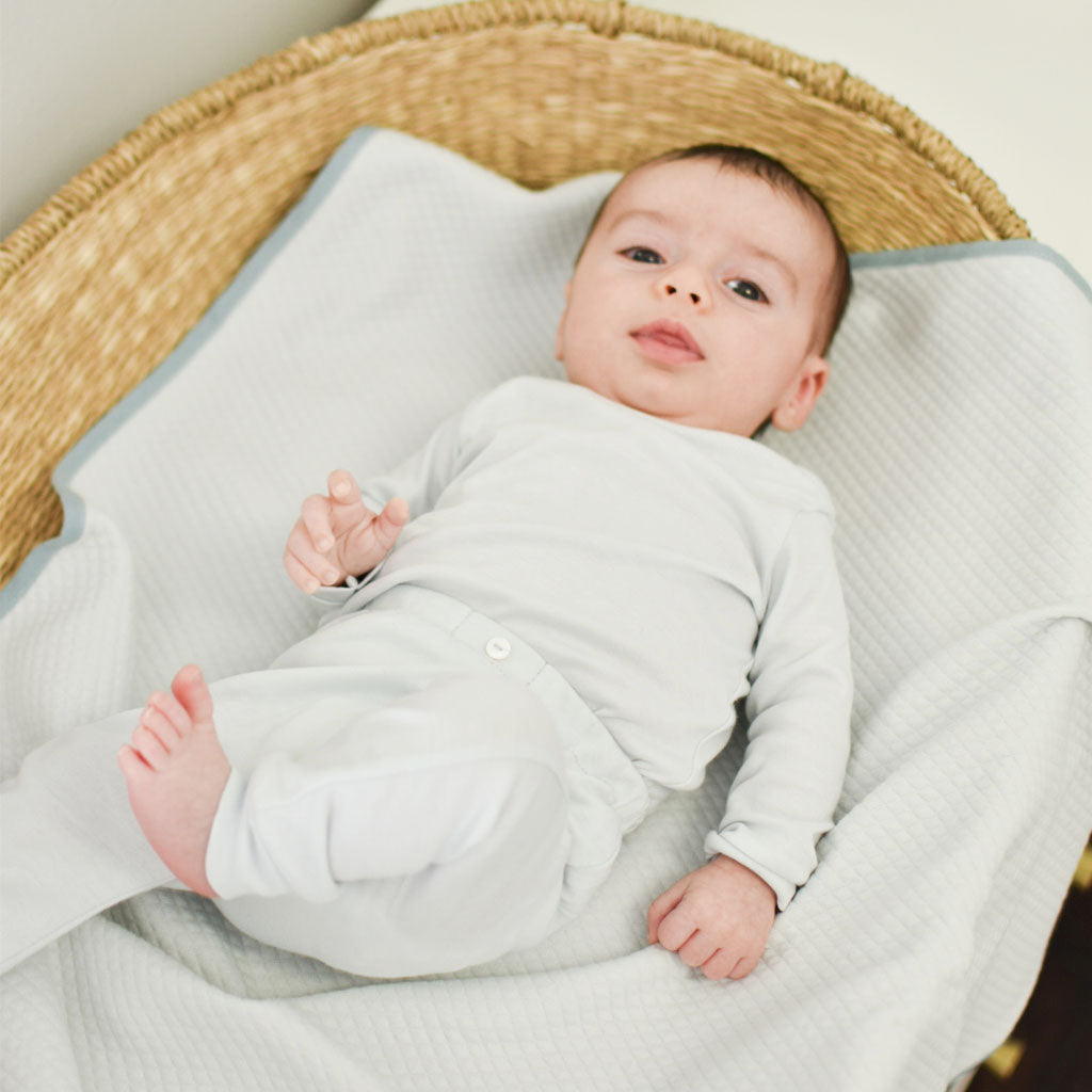 A baby dressed in Aiden Pima Top & Leggings, lying comfortably in a woven basket with a soft white liner, looking directly at the camera with a gentle expression.