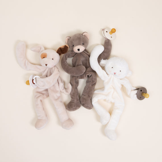 Three Silly Buddy Pacifier Holders, including a rabbit, a bear, and a lamb from Baby Beau & Belle, each holding a small wooden ring with a pacifier clip, arranged side by side on a light beige background.