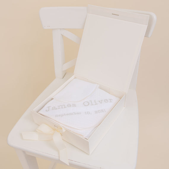 An elegant white Baby Boy Personalized Gift Set from Baby Beau & Belle with "James Oliver September 10, 2021" embroidered on fabric inside, placed on a white chair with a cream background.