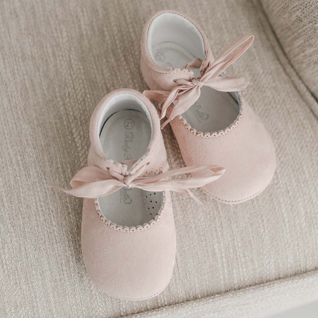 A pair of pink Elizabeth Suede Tie Mary Janes, perfect for a christening, placed neatly on a soft, beige fabric surface.