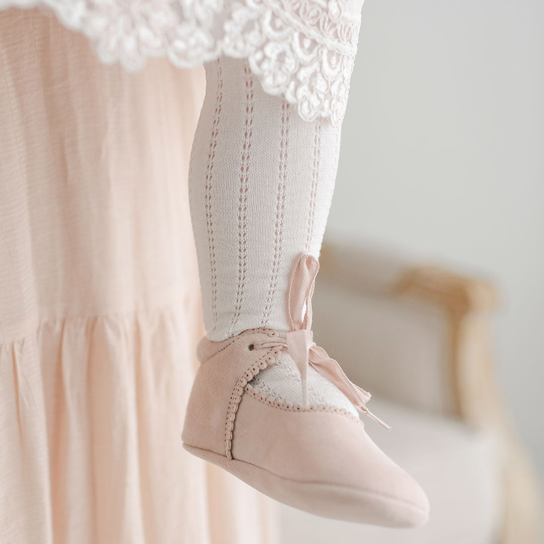 Close-up of a person's foot wearing a pair of Elizabeth Suede Tie Mary Janes, secured with a tied ribbon under a lace-trimmed pink christening dress hem. The leg is clad in.