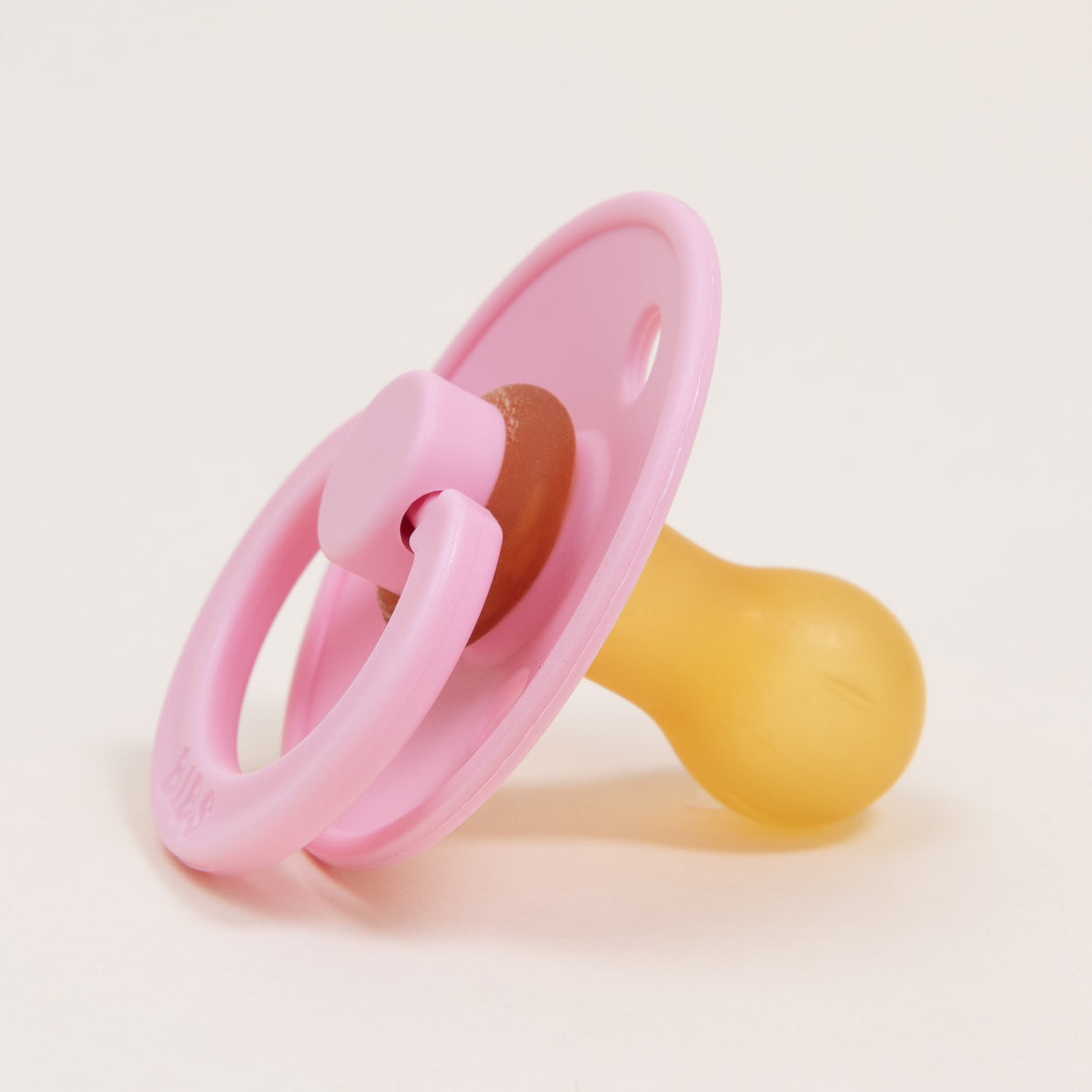 A Baby Pink Bibs Pacifier lying on a plain, light beige background. The pacifier has a prominent pink handle and shield with a yellow nipple.