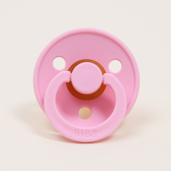 A Bibs Pacifier in Baby Pink, designed and manufactured in Denmark, with a circular rubber nipple and a round ventilation shield, labeled "bibs," on a light beige background.