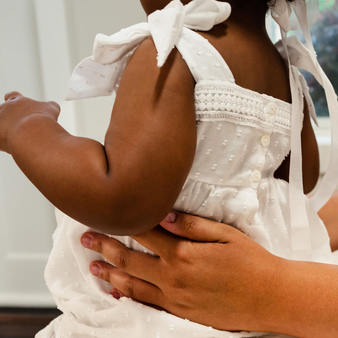 A close-up view of an adult's hands embracing a toddler wearing the Mila Cotton Gown. The focus is on the back button closures and  fabric details.