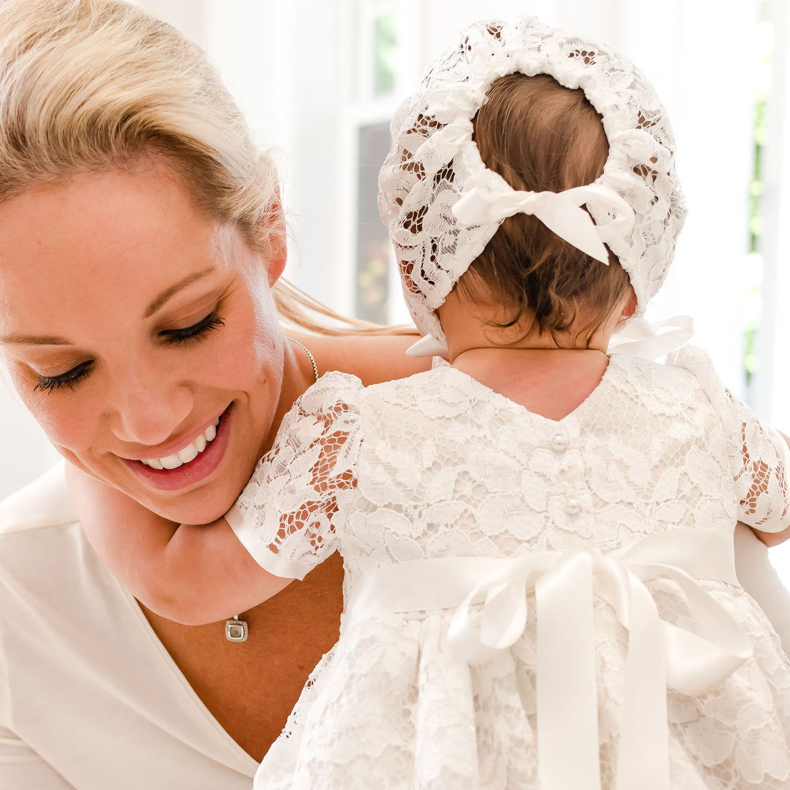 A joyful woman holding a baby girl in a traditional white Rose Lace Dress and matching Rose Lace Bonnet, both smiling in a brightly lit room.