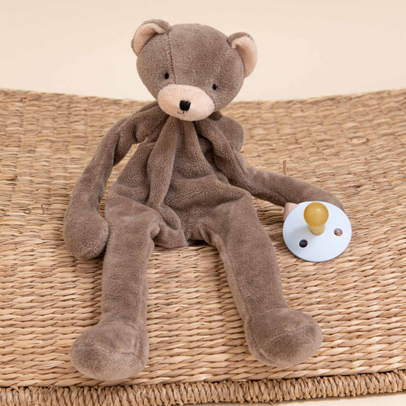 A soft, brown Austin Bear Buddy sitting on an upscale woven mat with a white and blue Pacifier Holder next to it. The background is neutral colored.