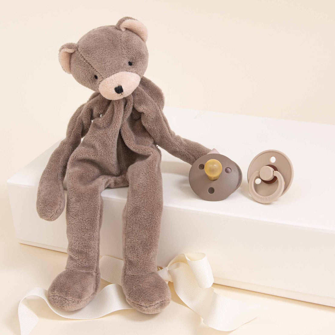 A plush Dylan teddy bear in a soft beige tone sits on a white block next to two pacifiers and a newborn outfit, one on a ribbon, against a creamy background.