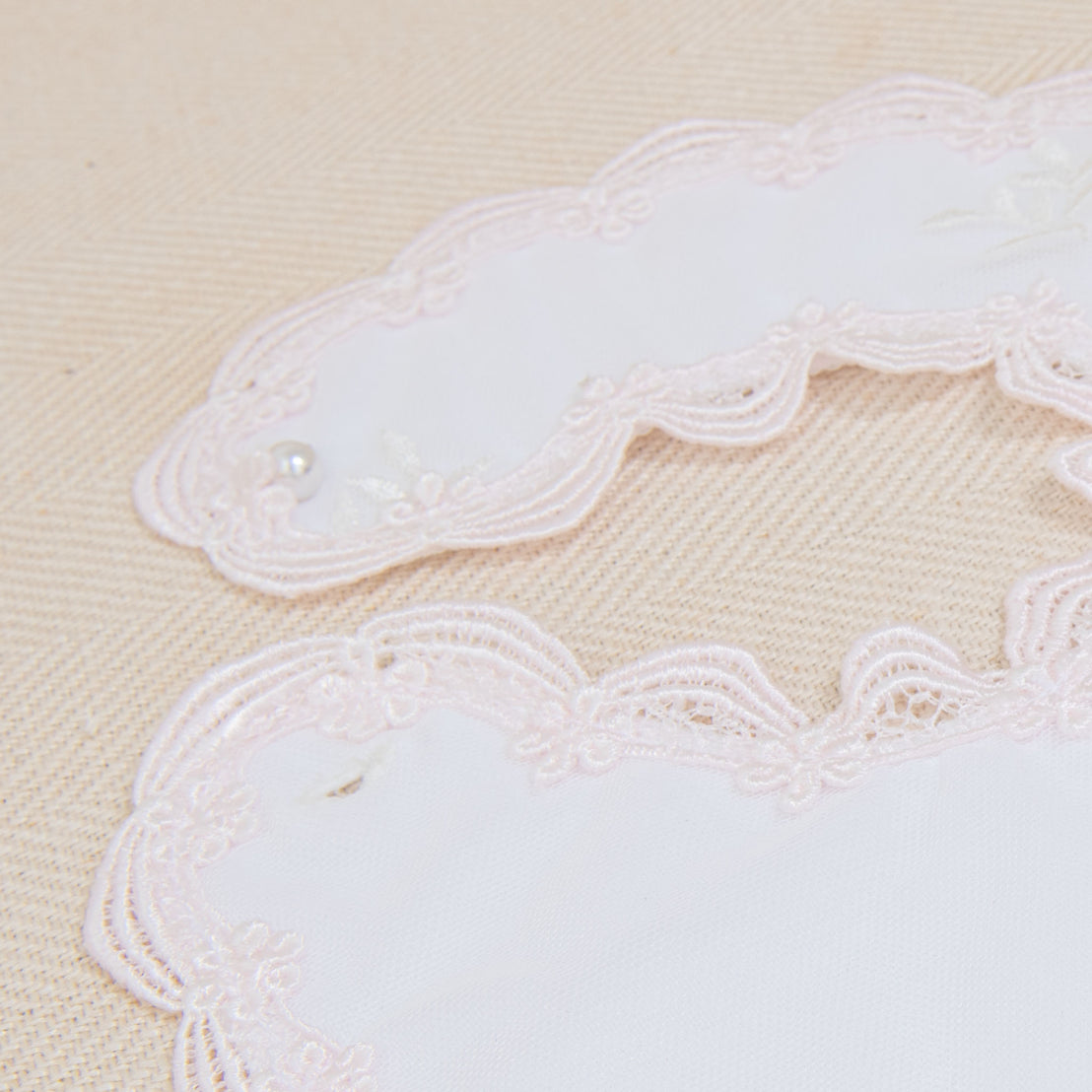 Close-up of delicate pink lace with vintage-inspired floral embroidery and tiny pearl embellishments on a beige fabric background, highlighting detailed craftsmanship of the Joli Bib.