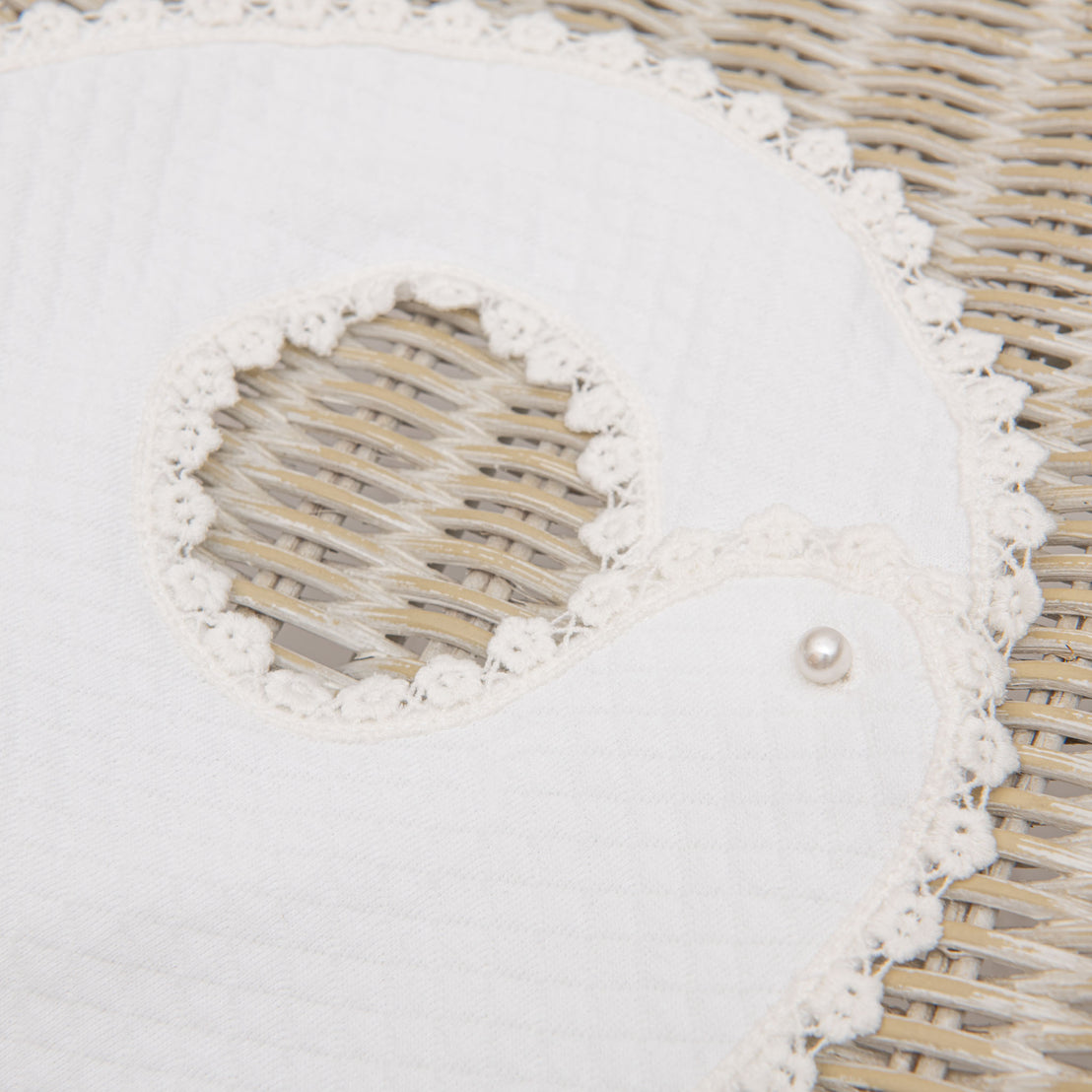 Close-up of a vintage, white, lacy Eliza Bib with a pearl button detail, placed on a woven chair. The fabric features delicate cut-out patterns.