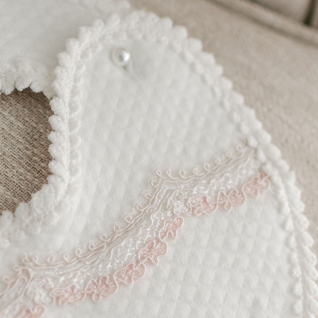Close-up of an upscale, Elizabeth Bib with delicate lace trim and an embroidered christening design, featuring a single button closure, displayed on a soft textured surface.