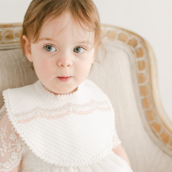 A toddler with striking blue eyes sits on an elegant chair, wearing a white Elizabeth Bib with a detailed lace collar, set against a soft focus on a soothing and light backdrop.