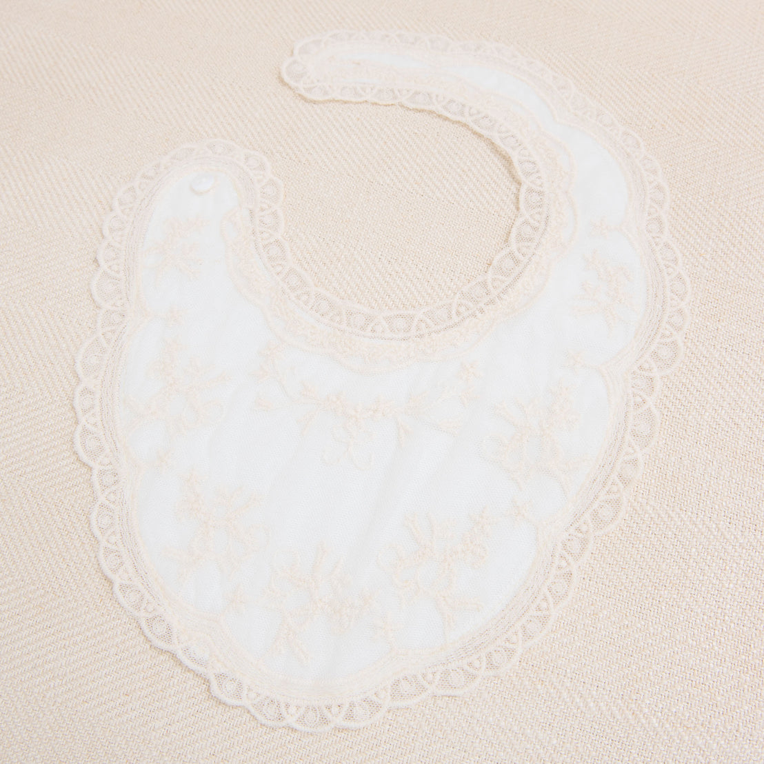 The Jessica Cotton Bib, featuring an intricate floral embroidery champagne lace over ivory cotton, displayed on a beige fabric background.