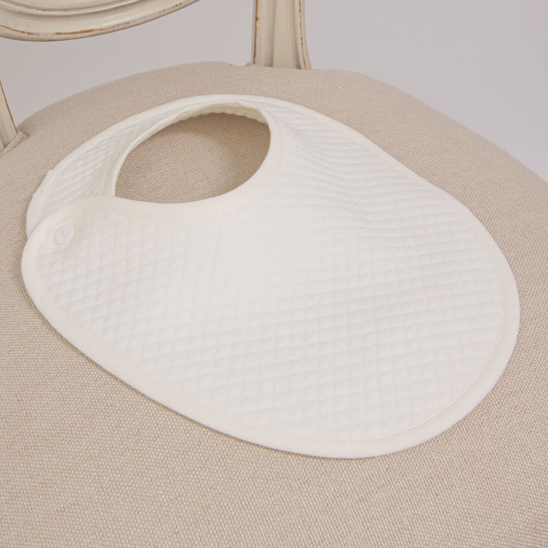 The Owen Bib crafted from ivory quilted cotton and a single button closure placed neatly on a vintage light beige upholstered chair.
