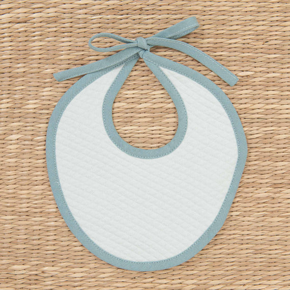 A Aiden Bib in a simple oval shape with a blue border and upscale ribbon, displayed flat on a textured, woven background.