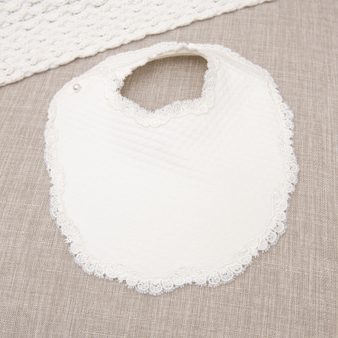 A white Madeline Bib with a scalloped lace trim and a single button closure, resting on a textured gray fabric surface.