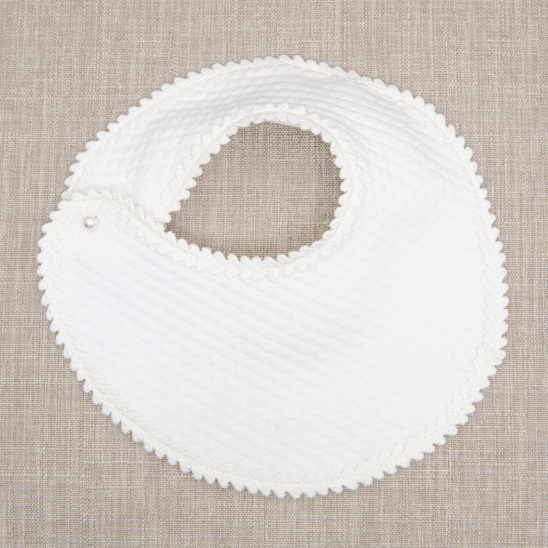 Flat lay photo of the Tessa Quilt Bib. The bib is made with a soft quilted cotton, accented with a floral edge lace, and features a pearl style button closure. 