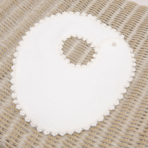 A white Eliza Bib with a decorative edge and a single pearl button, displayed on a textured wicker background, exudes an upscale vintage charm.