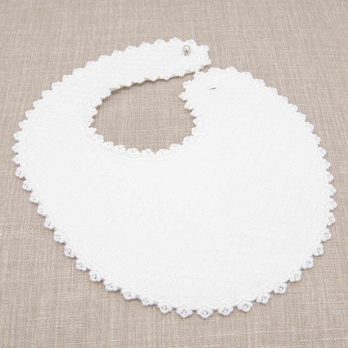A Mila bib made from white quilted cotton with lace-edged scallop design and a pearl style button closure. The bib is displayed on a beige fabric background.
