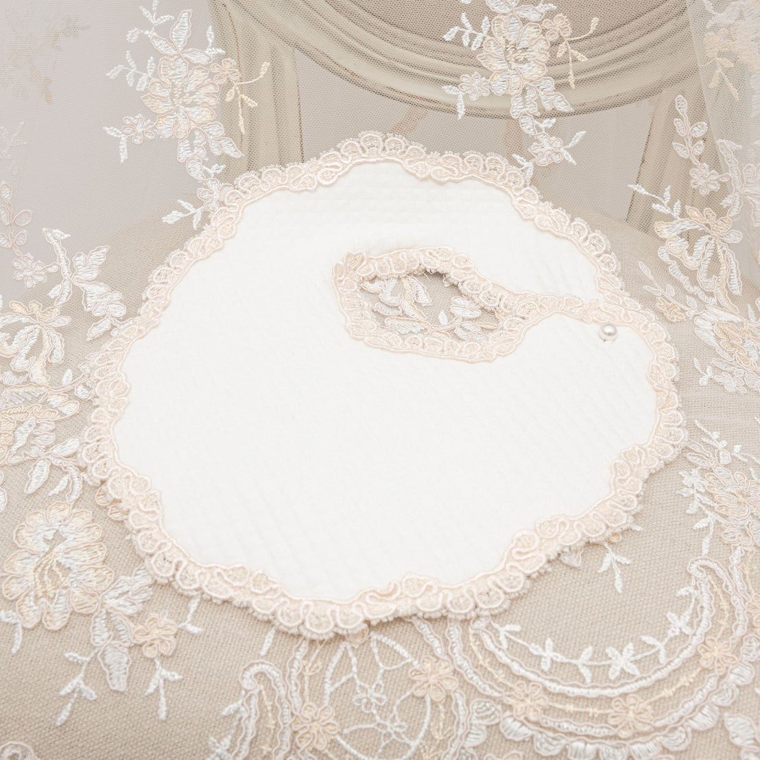 An elegant Kristina Bib placed on a vintage-inspired white embroidered lace fabric background, with floral patterns and a scalloped edge visible, creating a delicate and refined texture contrast.