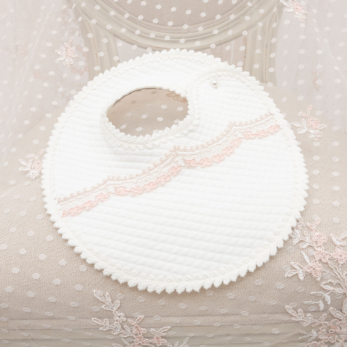 A white textured Elizabeth Bib with a pink embroidered design, displayed on a sheer fabric with floral details for christening or baptism. The bib features a snap button closure near the neckline. 