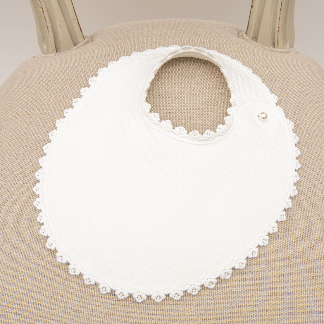 A close-up of a June Bib, displayed on a beige chair. The bib features a rounded neckline, a snap button closure, and a textured white quilted cotton material with delicate Venice lace trim.