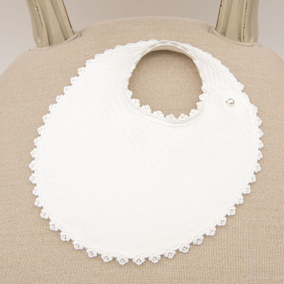 A close-up of a June Bib, displayed on a beige chair. The bib features a rounded neckline, a snap button closure, and a textured white quilted cotton material with delicate Venice lace trim.