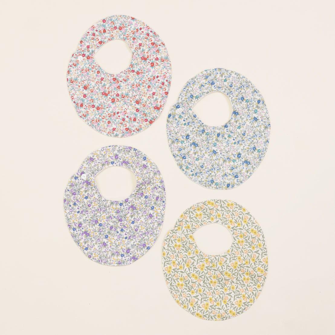 Four Petite Fleur Bibs, each featuring a central hole and a vintage-inspired, upscale disc with floral patterns, arranged in a two-by-two grid on a light background.