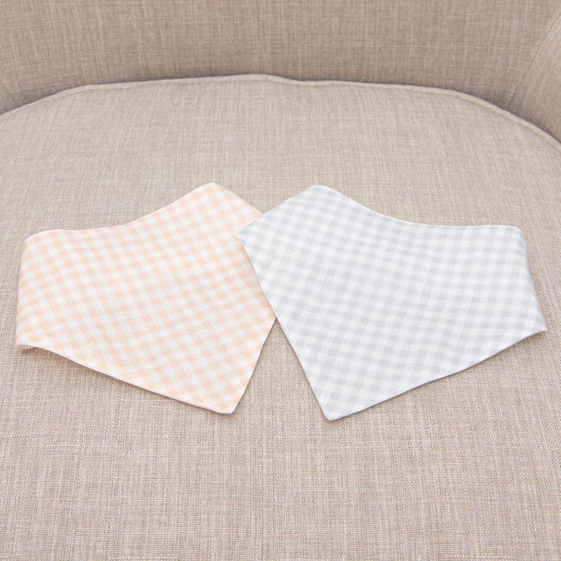 Two Ian Bandana Bibs with traditional checkered patterns, one peach and the other light blue, laid on a grey sofa.