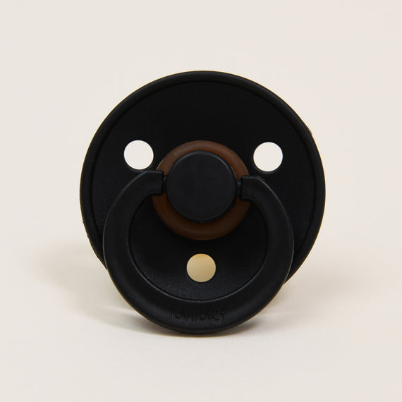 A black and brown Bibs Pacifier 2 Pack placed on a plain beige background, facing directly at the camera. The pacifier has a circular shield with three ventilation holes.