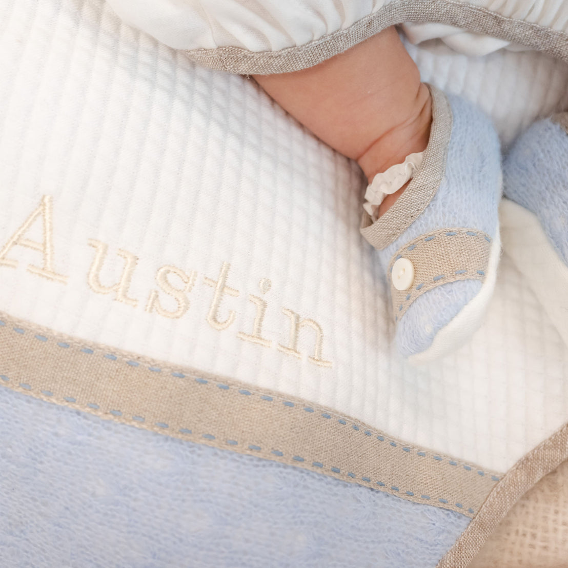 A close-up of a white and blue Austin Personalized Blanket with the name "Austin" embroidered in a delicate script, resting in a woven basket. A baby foot wearing baby booties is in the upper right corner.