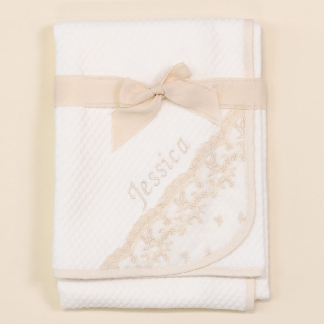 The Jessica Personalized Blanket with "Jessica" embroidered in champagne, adorned with matching lace trim on the corner. A champagne satin ribbon tied in a bow secures the blanket set against a beige background.