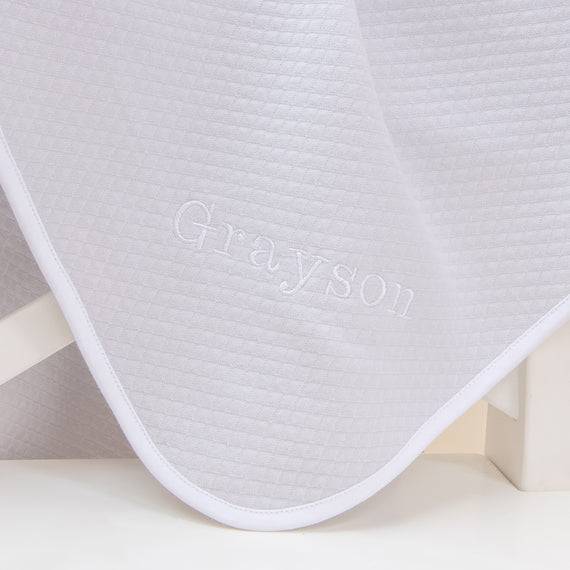 Flat lay photo of the Grayson Personalized Blanket. It is made from quilted textured cotton in grey with a white pima cotton trim. On the corner is the name "Grayson" embroidered in white thread.