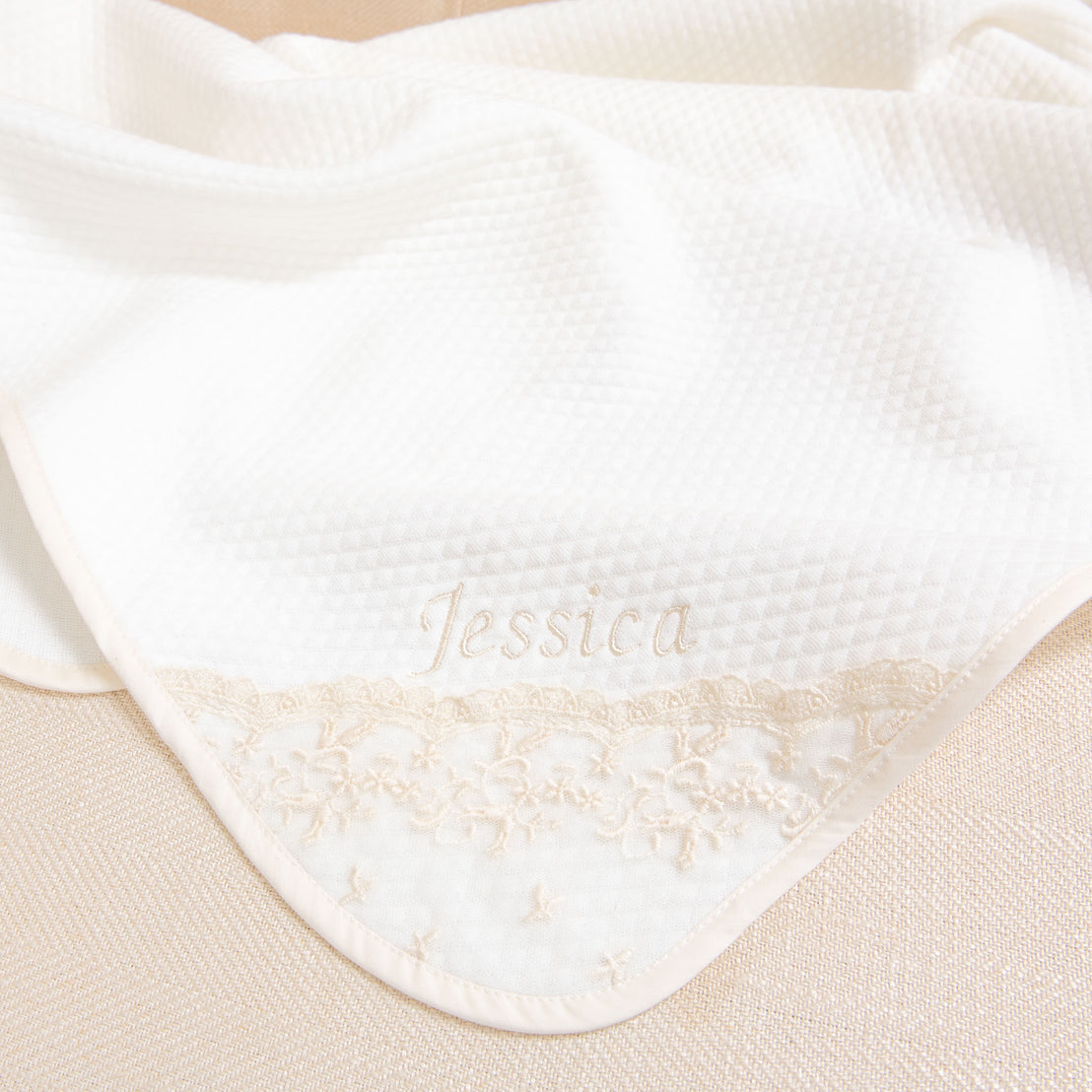 Close-up of the Jessica Personalized Blanket with the name "Jessica" embroidered in delicate script, adorned with a champagne lace trim and embroidered floral details, set against a soft beige background.