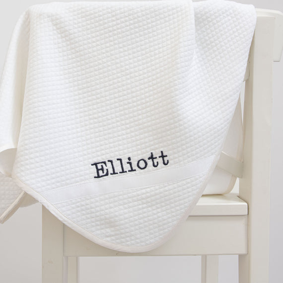 Flat lay photo of the Elliott Personalized Blanket, a soft white 100% quilted cotton, one corner is detailed with ribbon and the name "Elliott" embroidered in navy.
