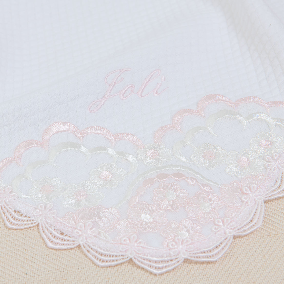 A close-up photo of a fabric with the embroidered name "Joli" in pink thread, partially overlaying an intricate white and pink lace design on a textured off-white background.